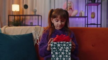 Toddler girl opening gift box with light glow inside, smiling joyfully, looking amazed satisfied with nice present, unexpected birthday surprise. Little child kid at home in evening living play room