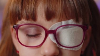 Close-up portrait of sick ill girl face looking at camera, having protective bandage plaster patch scratch on one eye. Injury, blind. Child kid in glasses. Medical eyesight vision therapy correction