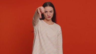 Dislike. Upset unhappy pretty woman in pullover showing thumbs down sign gesture, expressing discontent, disapproval, dissatisfied, dislike. Young adult girl. Indoor studio shot on red background