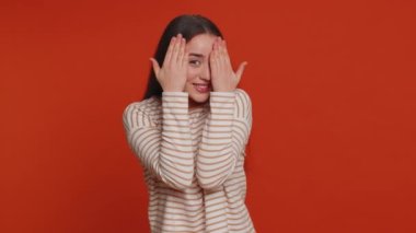 Nosy curious young woman closing eyes with hand and spying through fingers, hiding and peeping, binocular gesture, exploring way, seeking something in distance. Girl isolated on red studio background