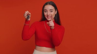 Pretty young woman in crop top real estate agent lifting hand showing the keys of new home house apartment, buying, renting property, mortgage loan. Millennial girl isolated on red studio background