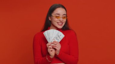 Rich pleased boss millennial woman waving money dollar cash banknotes bills like a fan, success business career, lottery winner, big income, wealth. Young pretty girl isolated on red background