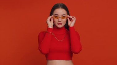 Portrait of happy pretty young woman in crop top wearing sunglasses, looking at camera with toothy charming smile, flirting, expressing optimism. Millennial girl isolated on red studio background