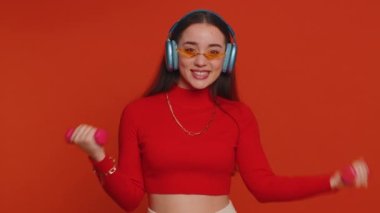 Young fit woman listening music via headphones, working out, pumping up arm muscles lifting pink dumbbells, practicing pilates, healthcare. Pretty girl indoors isolated alone on studio red background