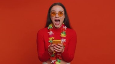 Cheerful tourist woman with hawaiian lei in sunglasses using smartphone celebrating winning holiday resort vacation tickets pass. Tourism, summer trip to sea. Happy pretty girl on red wall background