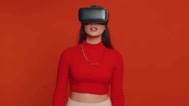 Lovely woman using headset helmet to play simulation game app online. Watching virtual reality 3D 360 video content. One girl in VR goggles isolated on studio red background. Futuristic technology