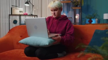 Young woman with short blonde hair freelancer at home night room sitting on couch, opens laptop start working. Girl works on notebook, sends messages, makes online purchases, watching movies, working