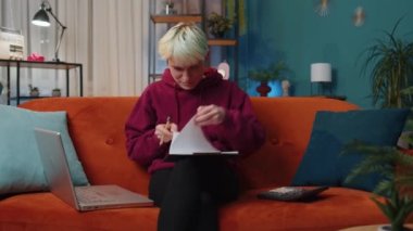 Young woman accountant working with financial graph documents, calculate taxes for charts and graph reports of home income on couch. Businesswoman working on laptop computer planning expenses budget
