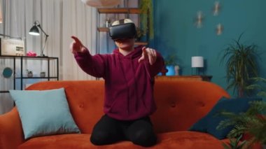 Young woman using virtual reality futuristic technology VR app headset helmet to play simulation 3D 360 video game, watching film movie at modern home apartment. Blonde girl in goggles sitting on sofa