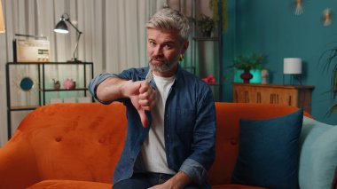 Dislike. Upset middle-aged man showing thumbs down sign gesture, expressing discontent, disapproval, dissatisfied bad work at modern home apartment indoors. Displeased old guy in living room on sofa