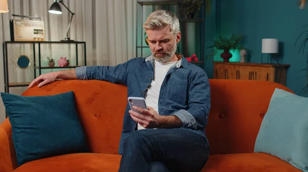 Bearded middle-aged man sitting on sofa uses mobile phone smiles at night home apartment. Mature guy texting share messages content on smartphone social media applications online, watching relax movie