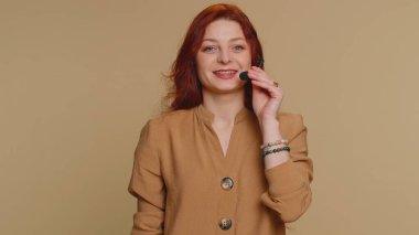Gülümseyen kızıl saçlı kadın, kulaklık takan, serbest çalışan, çağrı merkezi ya da destek operatörü yardım hattı, müşteriyle ya da meslektaşıyla konuşan, iletişim desteği olan. Bej arka planda genç bir kız