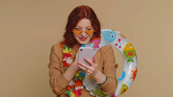 Cheerful young tourist woman with luggage in sunglasses with smartphone celebrating winning holiday resort vacation tickets pass. Tourism, summer trip to sea. Happy redhead girl on beige background
