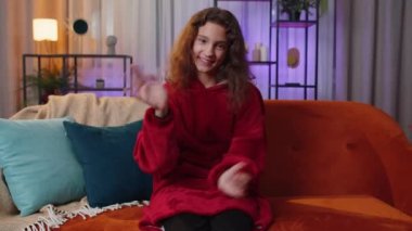 Preteen girl smiling friendly at camera and waving hands gesturing hello, hi, greeting or goodbye, welcoming with hospitable expression. Child kid at home in night evening living play room on couch