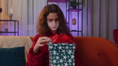 Preteen girl opening gift box with light glow inside, smiling joyfully, looking amazed satisfied with nice present, unexpected birthday surprise. Young child kid at home in evening living play room