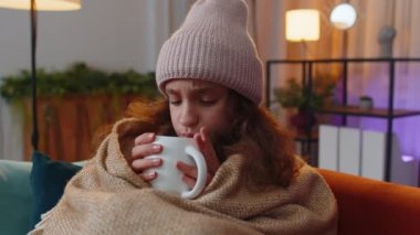 Sick preteen girl wear hat wrapped in plaid sit alone shivering from cold on sofa drinking hot tea in unheated apartment without heating due debt. Unhealthy child feeling discomfort try to warming up