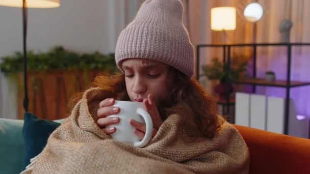病気の前の女の子は 有料で包まれた帽子をかぶるだけで 借金を加熱することなく 加熱されていないアパートで熱いお茶を飲むソファの寒さから震えて座っています 不健康な子供は不快感を感じて温暖化しようとする — ストック動画