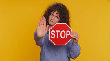 Serious curly haired woman say No, hold inscription text red stop danger sign, warning of finish, prohibited access, declining communication, body language, trouble protest. Girl on yellow background