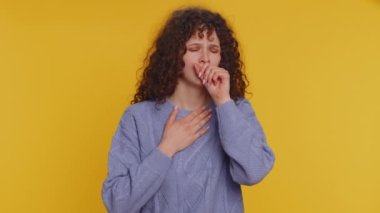 Unhealthy curly haired woman coughing covering mouth with hand, feeling sick, allergy, viral infection symptoms, coronavirus pandemic. Young adult teen girl isolated alone on yellow studio background