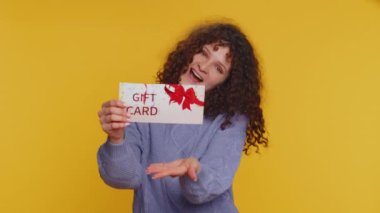 Young woman showing pointing on gift discount certificate voucher coupon for store holidays sale. Curly haired one girl with shopping christmas surprise card isolated alone on yellow studio background