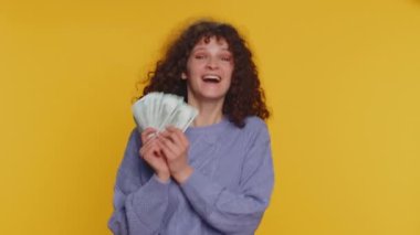 Rich pleased boss millennial woman waving money dollar cash banknotes bills like a fan, success business career, lottery winner big income wealth. Young curly haired girl isolated on yellow background