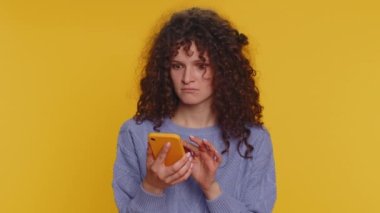 Hipster curly haired woman use mobile cell phone typing browsing, loses becoming surprised sudden lottery results, bad fortune, loss, fail. Young girl isolated alone on yellow studio wall background