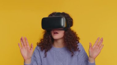 Young woman using headset helmet to play simulation game app online. Watching virtual reality 3D 360 video content. Girl in VR goggles isolated on studio yellow background. Futuristic technology