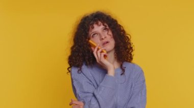 Curly haired woman tired sleepy freelancer talking on mobile phone with friend, making online conversation. Disinterested sad girl having annoyed boring talk on smartphone on yellow studio background