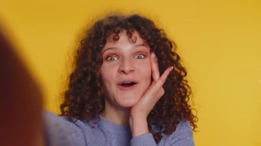 Oh my God, Wow. POV selfie portrait of excited amazed winner woman raising hands in surprise looking at camera with big eyes, shocked by sudden victory. Sincere curly haired girl on yellow background