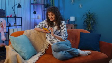 Kanepede oturan kıvırcık saçlı kadın gece evinde cep telefonu gülümsemesi kullanıyor. Çevrimiçi akıllı telefon sosyal medya uygulamaları üzerinden mesajları paylaşan, rahatlamış film izleyen bir kız