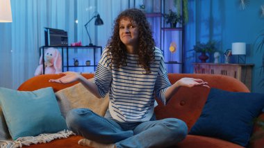 What. Why. Young woman raising hands in indignant expression asking reason of failure demonstrating disbelief irritation by troubles, quarrel at home apartment. Girl in evening room sitting on sofa