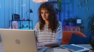 Young woman with curly hairstyle freelancer at home night room sitting at desk, opens laptop start working. Girl works on notebook, sends messages, makes online purchases, watching movies, working