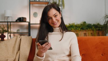 Brunette young woman sitting on sofa uses mobile phone smiles at home living room apartment. Girl texting share messages content on smartphone social media applications online, watching relax movie