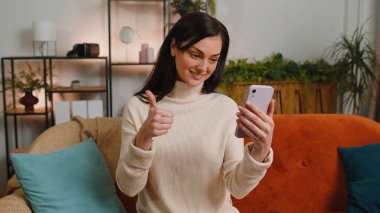 Young woman making smartphone call online video webcam conversation with friends sitting on couch at home in living room apartment. Happy excited girl enjoying mobile loudspeaker talking indoors