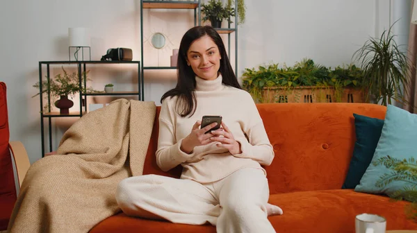 Young woman sitting on sofa uses mobile phone smiles at home living room apartment. Brunette girl texting share messages content on smartphone social media applications online, watching relax movie