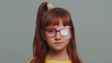 Sick ill preteen girl looking at camera having protective bandage plaster patch scratch on one eye. Injury, trauma, blind. Child kid wearing glasses at gray background. Medical eyesight vision therapy
