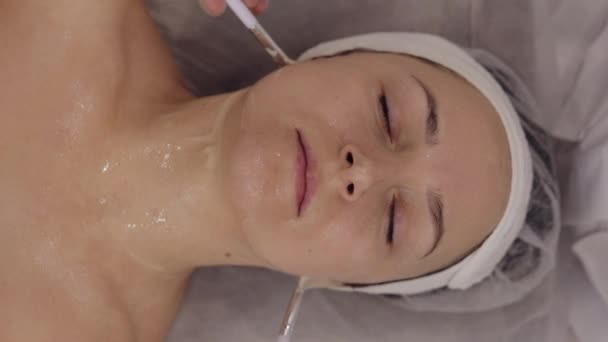 酶的治疗 美容师在中年妇女脸上用刷子涂润肤药用剥皮膏面膜 Spa上的女孩皮肤护理程序 面部美容术治疗 垂直视频拍摄 — 图库视频影像