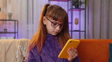 Toddler young girl use smartphone typing browsing, loses becoming surprised sudden lottery results, bad news, fortune loss, play game fail. Teen child kid at home in evening room sits on orange sofa