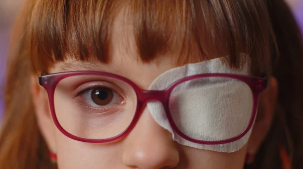 Close-up portrait of sick ill girl face looking at camera, having protective bandage plaster patch scratch on one eye. Injury, blind. Child kid in glasses. Medical eyesight vision therapy correction