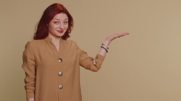 ブラウスで素敵な女性が親指を表示し 空の場所を指す 商業テキストの広告エリア 商品のプロモーションのためのスペースをコピーします 若い赤毛の女の子 室内スタジオショット上のベージュの壁の背景 — ストック動画