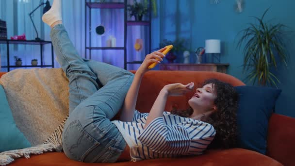 Young Woman Curly Hairstyle Lying Sofa Uses Mobile Phone Smiles — Video Stock