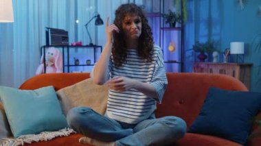 What. Why. Young woman raising hands in indignant expression asking reason of failure demonstrating disbelief irritation by troubles, quarrel at home apartment. Girl in evening room sitting on sofa