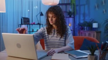 Young woman with curly hairstyle freelancer at home night room sitting at desk, opens laptop start working. Girl works on notebook, sends messages, makes online purchases, watching movies, working