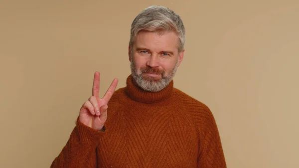 Middle-aged old man in sweater showing victory v sign, hoping for success and win, doing peace gesture, smiling with kind optimistic expression. Senior adult guy isolated on beige studio background