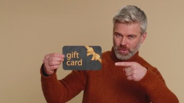 Senior mature man showing pointing on gift discount certificate voucher coupon for store holidays sale. Middle-aged guy with shopping christmas surprise card isolated alone on beige studio background