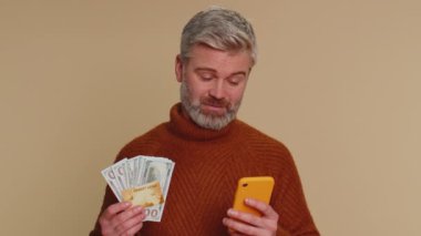 Sincere mature man customer using credit bank card and smartphone while transferring money, purchases online shopping, payment. Finance and internet. Senior old guy isolated alone on beige background