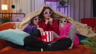 Excited teenage girl and little toddler child sisters under plaid eating popcorn and watching interesting tv serial, game, film, online social media movie. Three siblings children kids friends at home