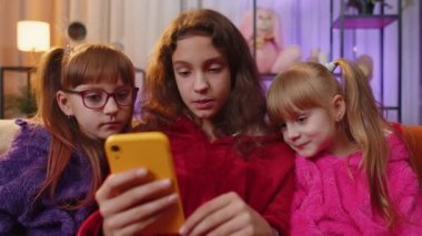 Teen girl, toddler child sisters using mobile phone texting share messages content on smartphone social media applications online, watching relax movie. Three siblings children kids at home playroom