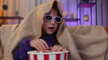 Excited toddler girl in 3D glasses eating popcorn and watching interesting tv serial, sport game, film, online social media movie content. Teen child kid at home in evening living play room on couch