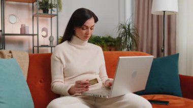 Young woman using credit bank card and laptop computer while transferring money, purchases online shopping, order food delivery at home apartment. Adult girl in day living room sitting on couch indoor